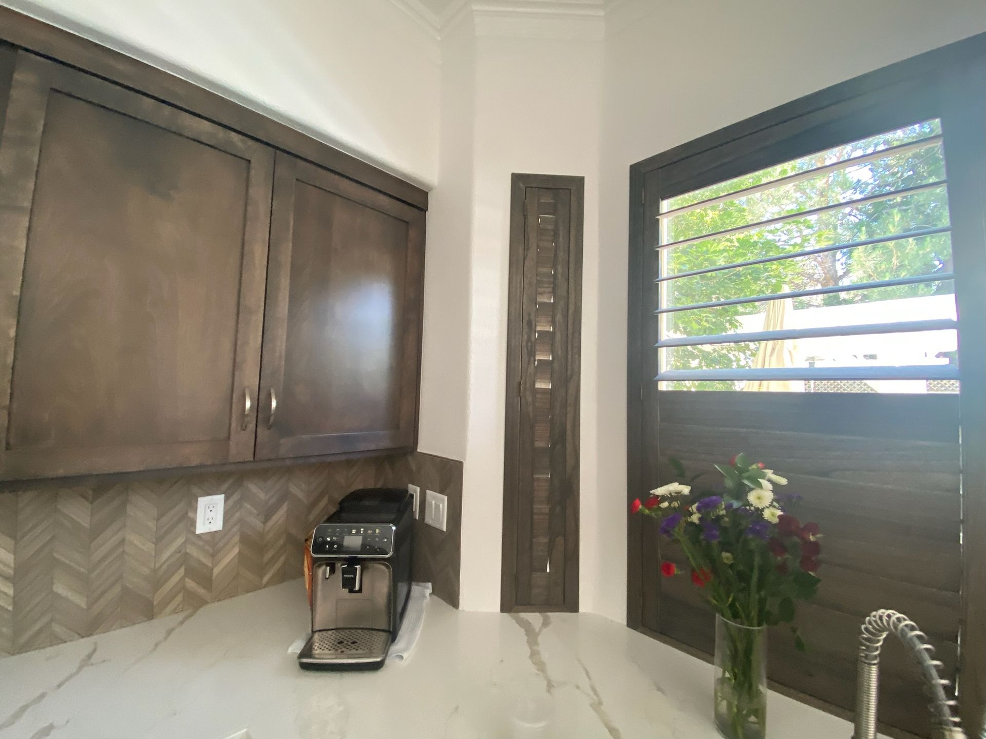 Modern kitchen with dark wood cabinets, coffee maker, sink, and vase of flowers by a window with blinds