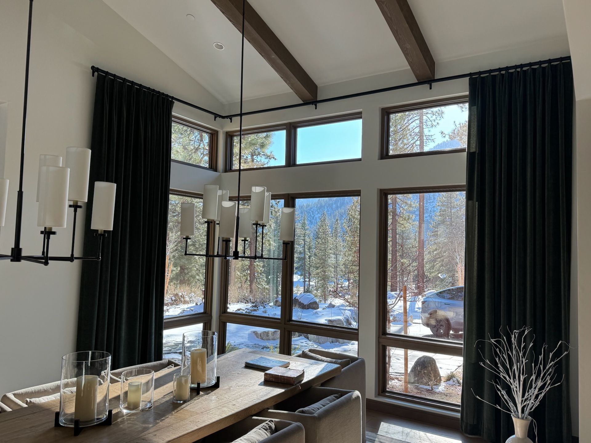 Bright dining room with tall windows, dark curtains, exposed beams, and a snowy view outside