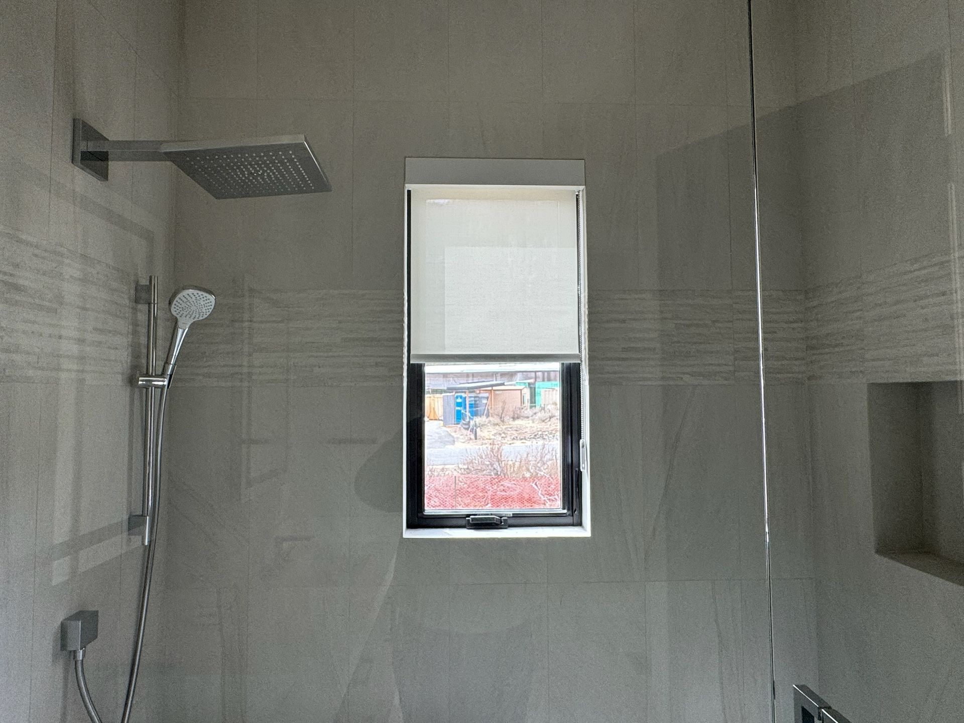 Modern tiled bathroom shower with rain showerhead, handheld sprayer, and a small window with a blind.