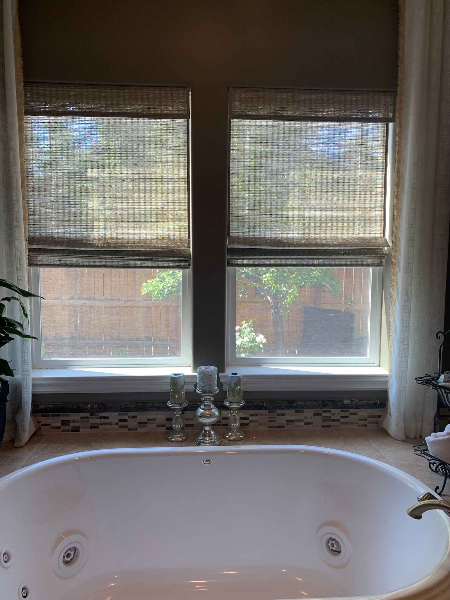 Bathroom with a white bathtub below a window with woven blinds and a row of small figurines on the sill