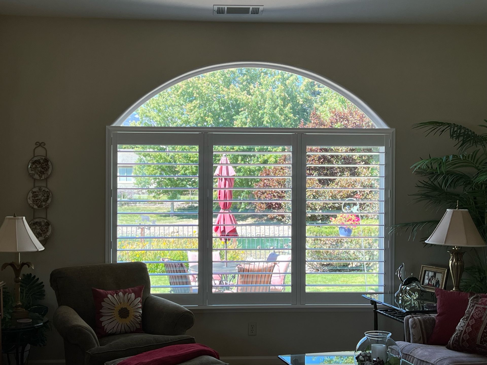 Living room with large arched window, white shutters, and patio view, flanked by lamps and chairs
