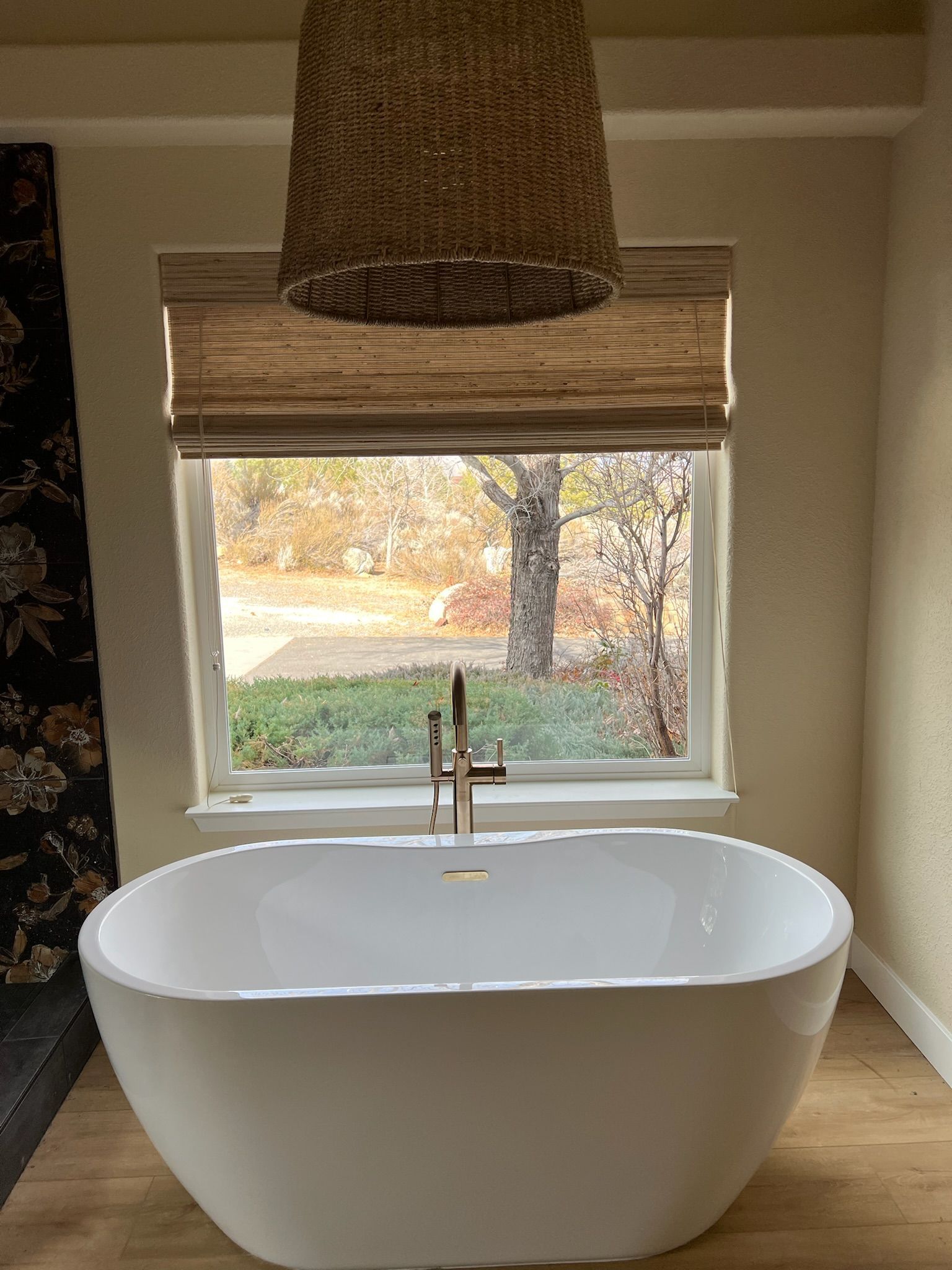 White oval bathtub under a window with a woven shade in a beige bathroom
