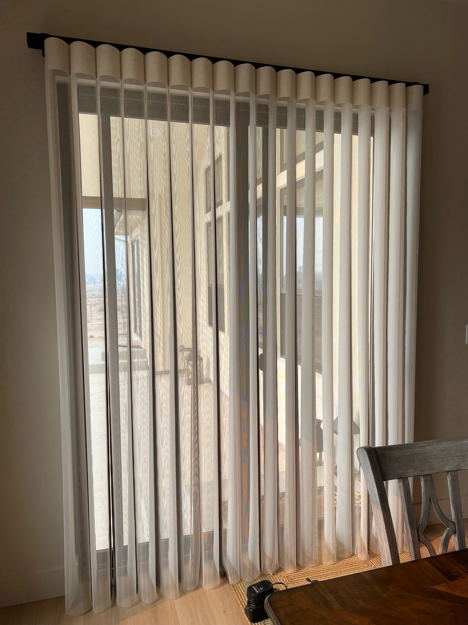 Sheer white curtains filtering sunlight in a living room near a wooden table and chair.