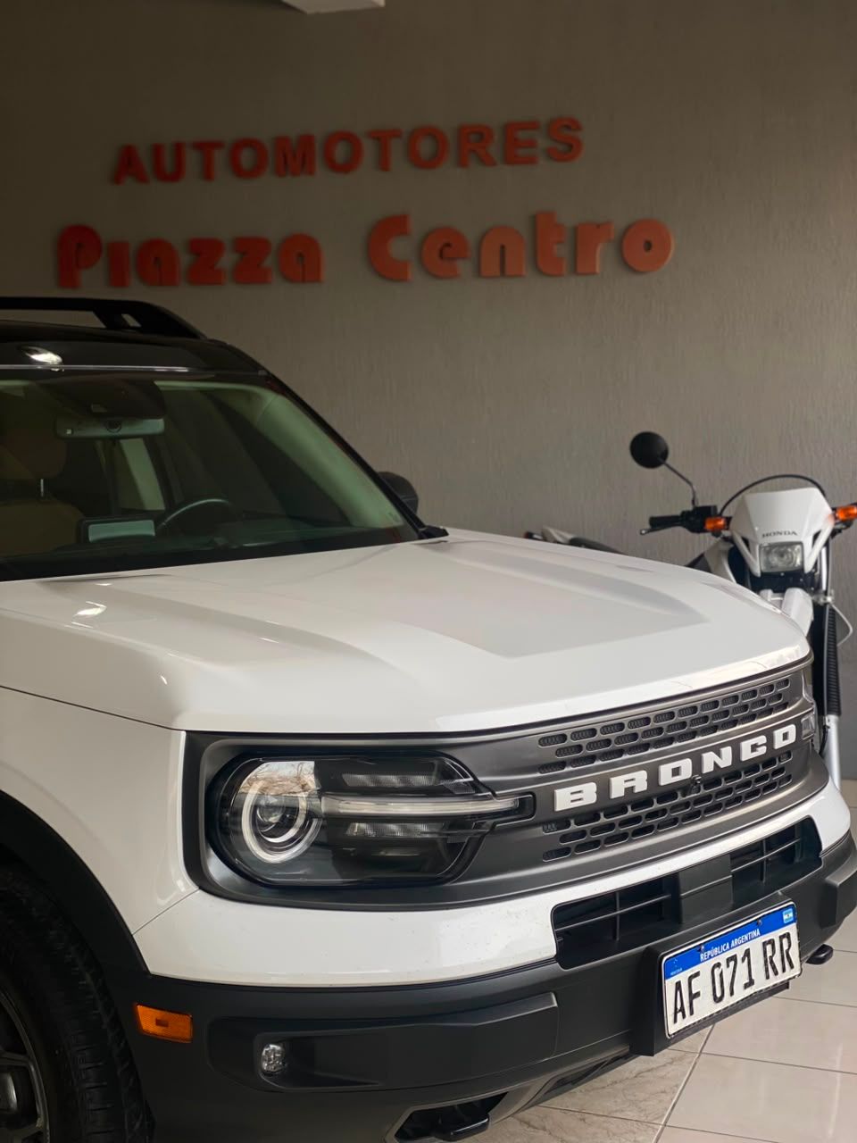 Un Ford Bronco blanco estacionado frente al letrero de Autotores Plaza Centro, con una motocicleta al lado.