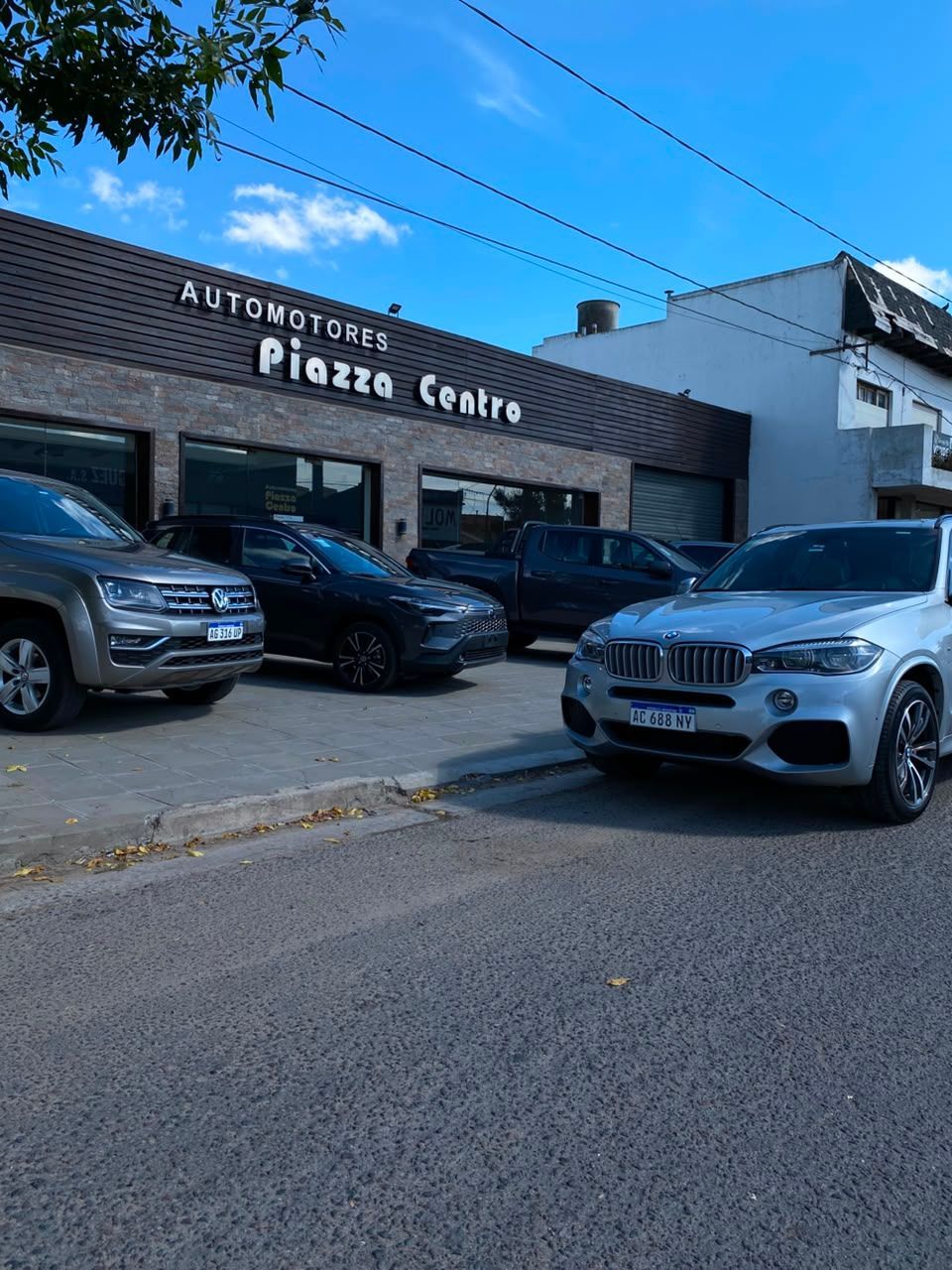 Aparcamiento exterior del edificio Alexander's Piazza Centro con varios coches aparcados bajo un cielo azul.