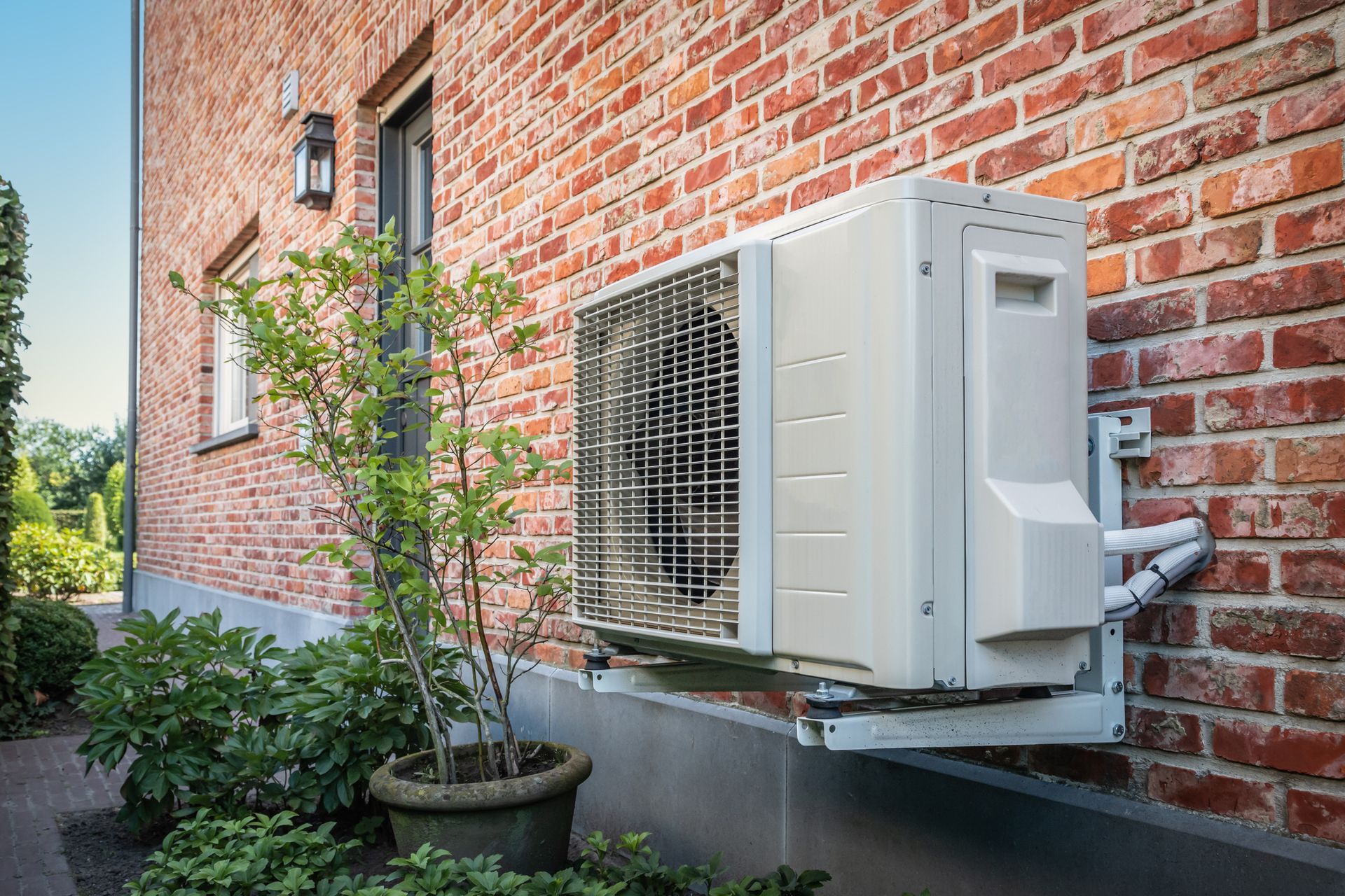 Air conditioning unit mounted on a red brick wall, next to a door and plants.