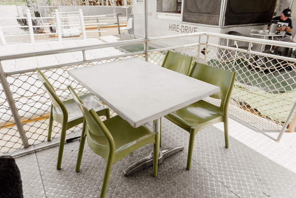 A table and chairs on a metal patio. The green chairs surround a white table. A person sits at another table.