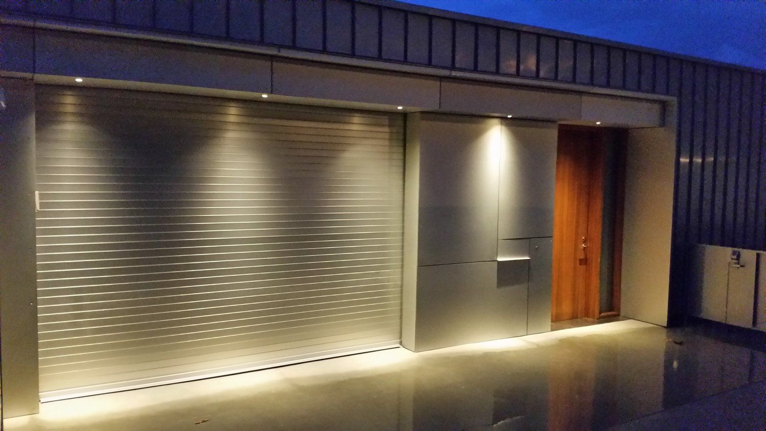 A stainless steel garage door with a wooden door is lit up at night.