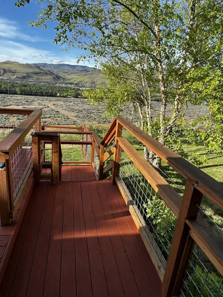 Wooden deck stairs with railings overlooking green fields, hills, and a tree under a bright sky.