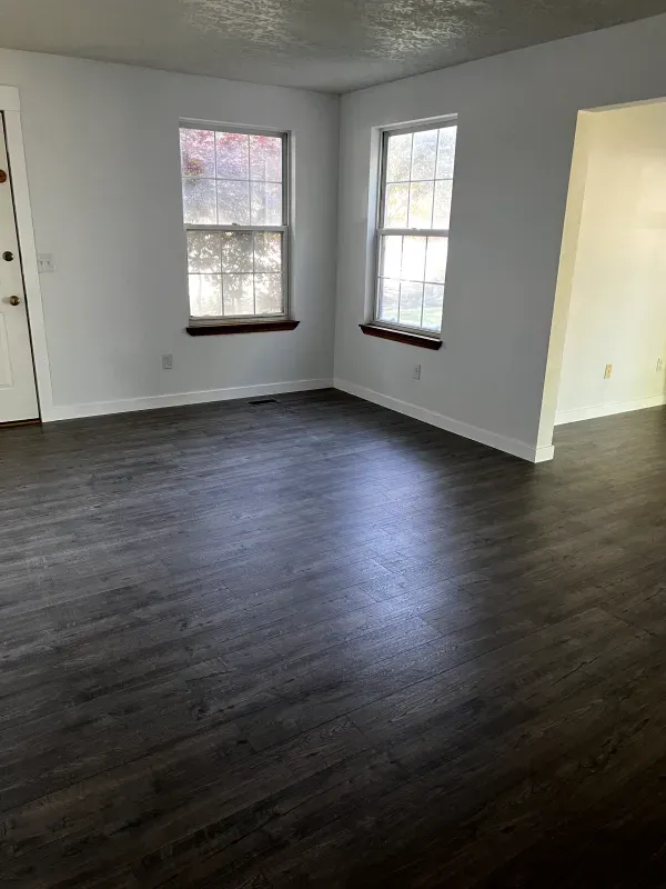 Empty room with dark wood floors, white walls, and two windows with dark trim.