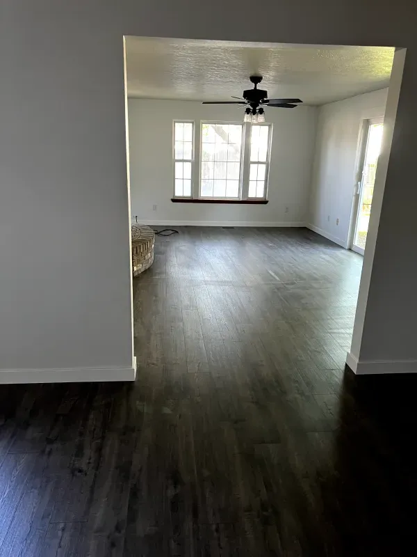 Empty living room with dark wood floors, ceiling fan, and large front window.