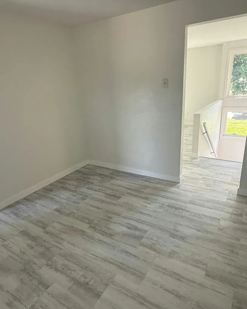Empty room with white walls, light wood floor, and doorway to a bright stairwell.