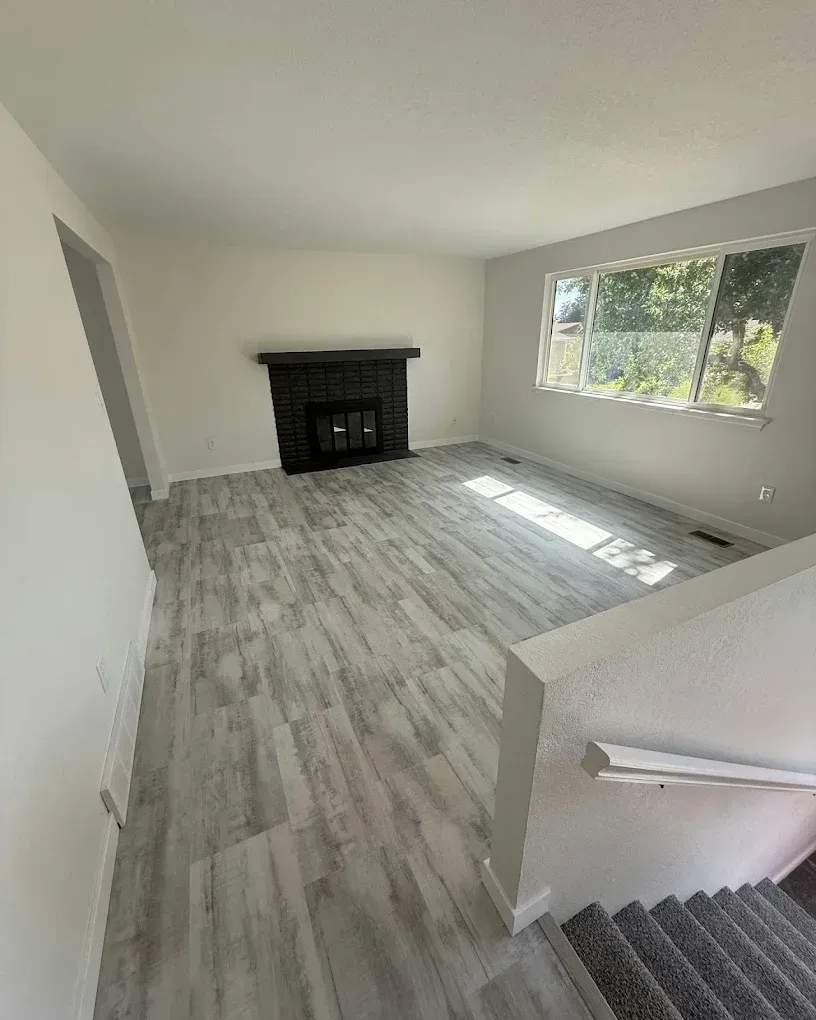 Empty living room with gray wood floors, black fireplace, and large sunlit window.