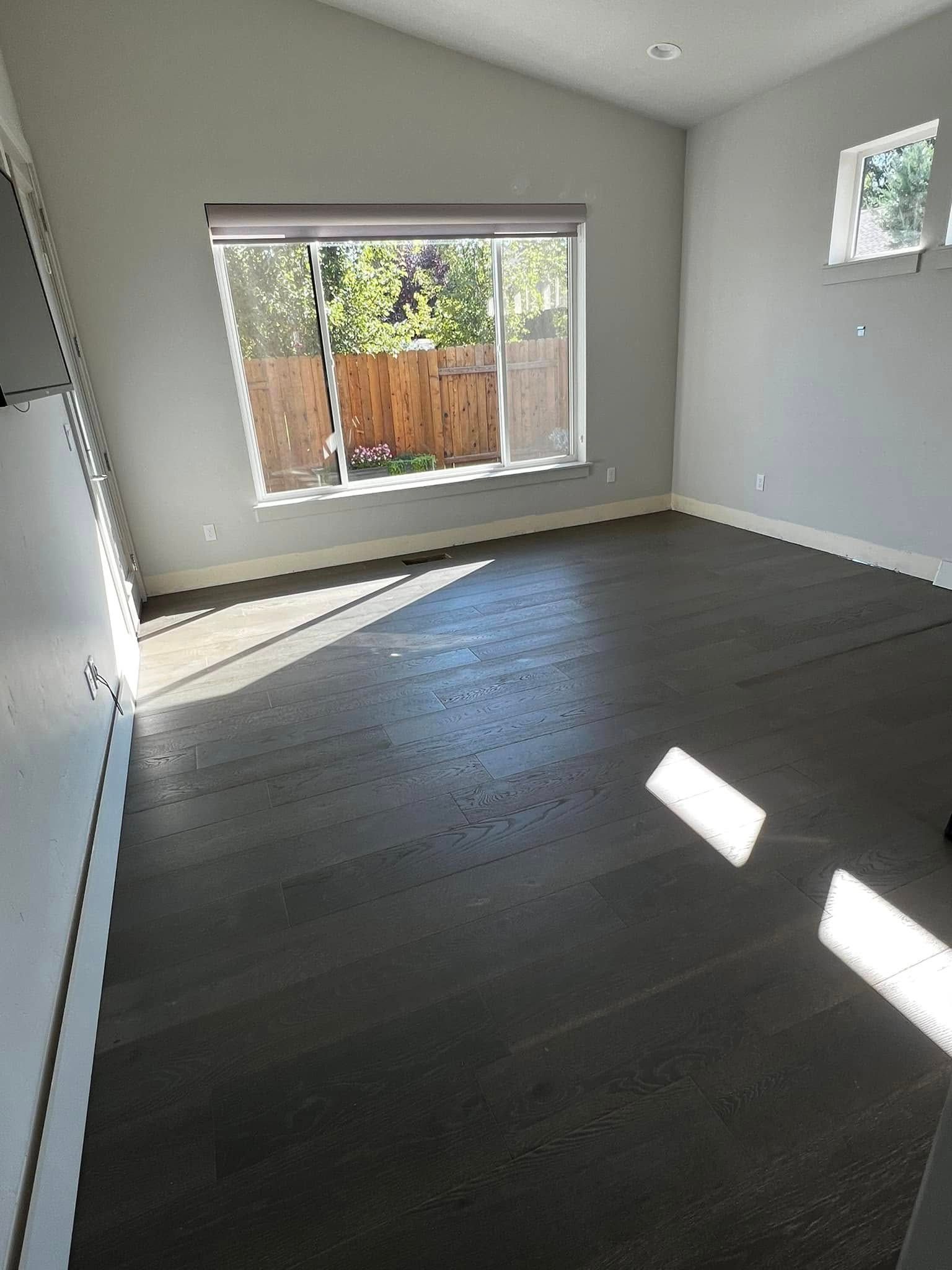 Empty gray room with dark wood floor, large window, and sunlight on the floor.