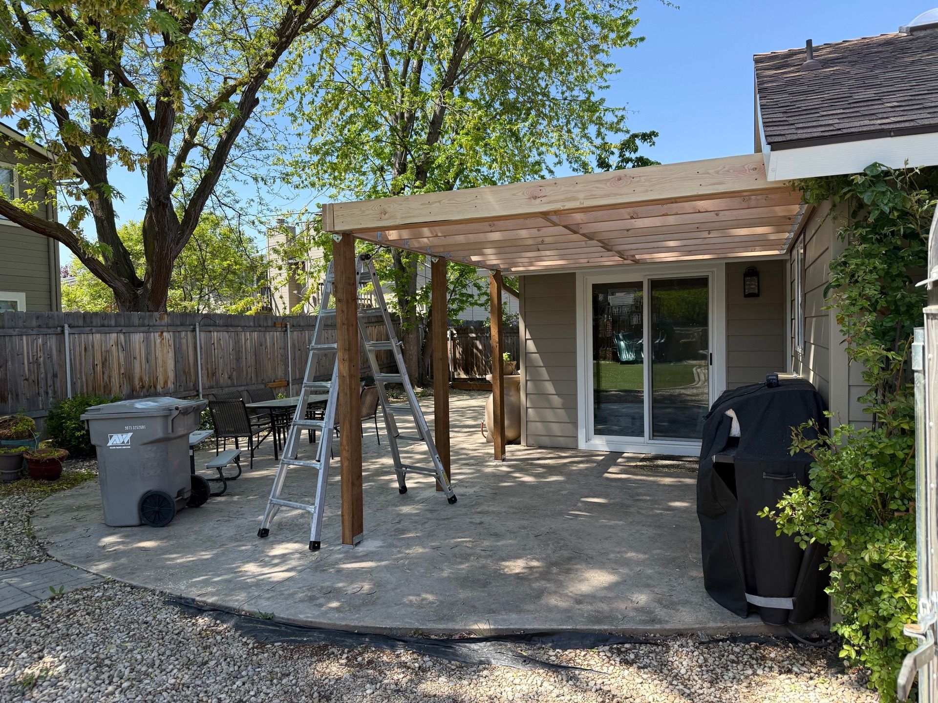 Backyard patio with a wooden pergola, ladder, trash bin, and barbecue grill beside a house.