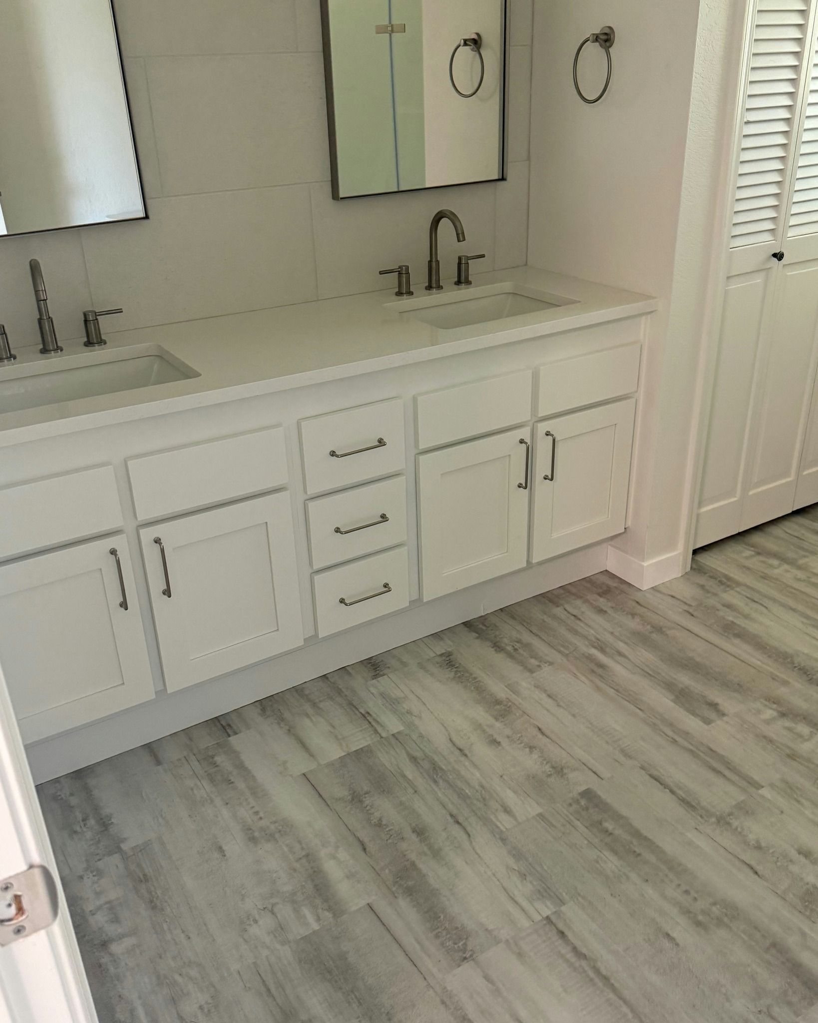 White bathroom vanity with mirror, double sinks, and light wood-look flooring beside a door.