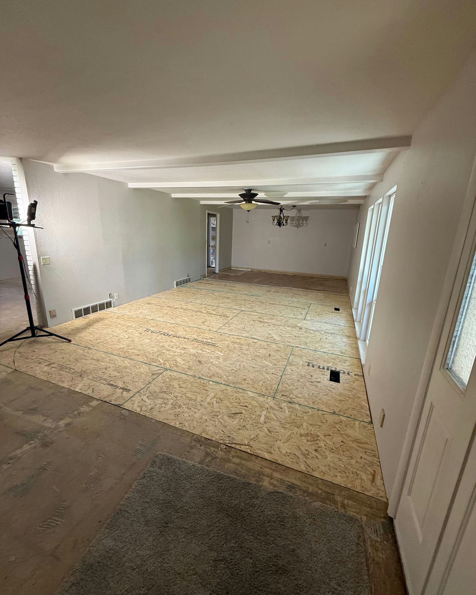 Empty living room with bare subfloor, white walls, ceiling fan, and window light.