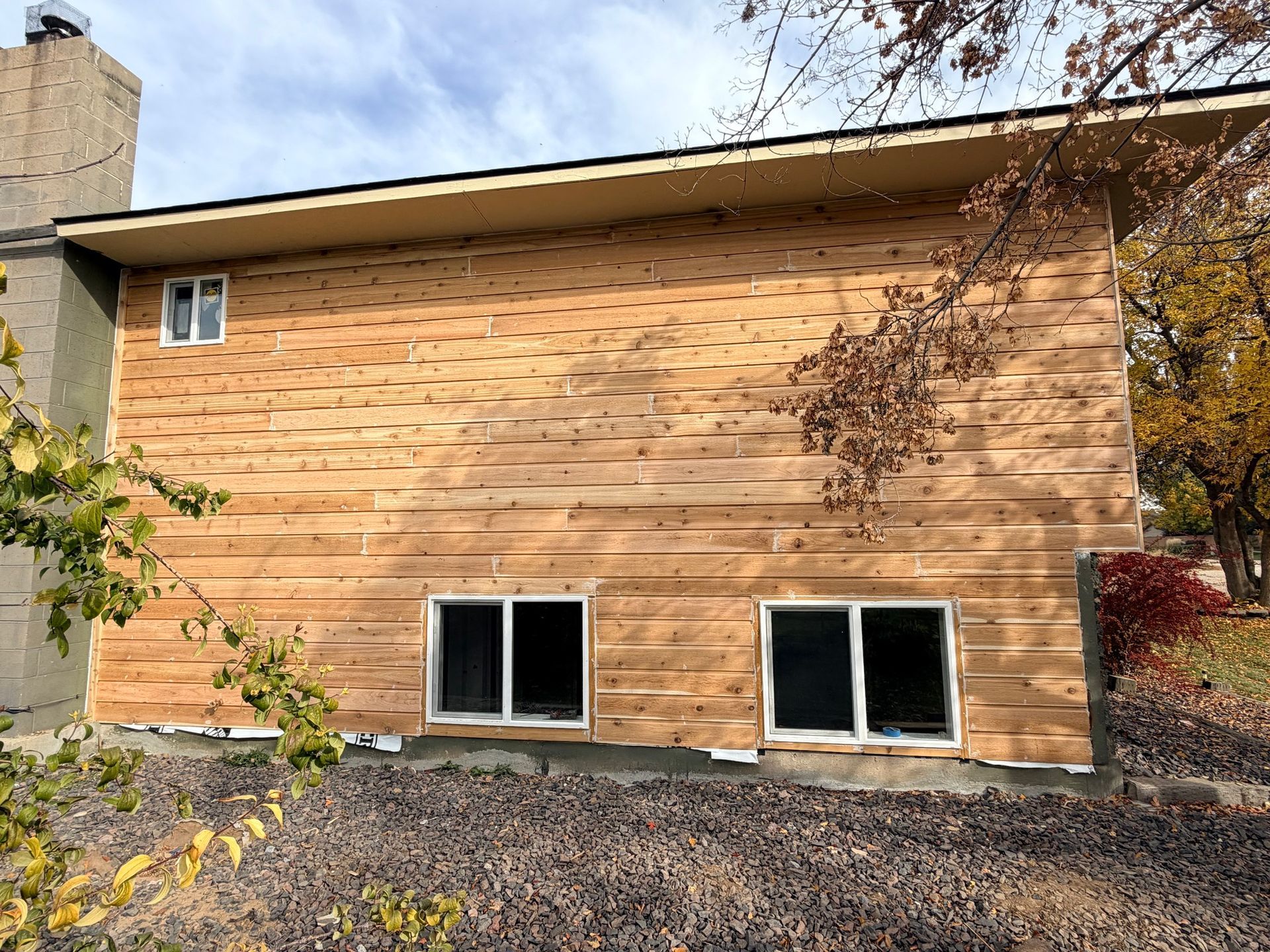 Wood-sided house exterior with a small window and two basement windows on a gravel yard