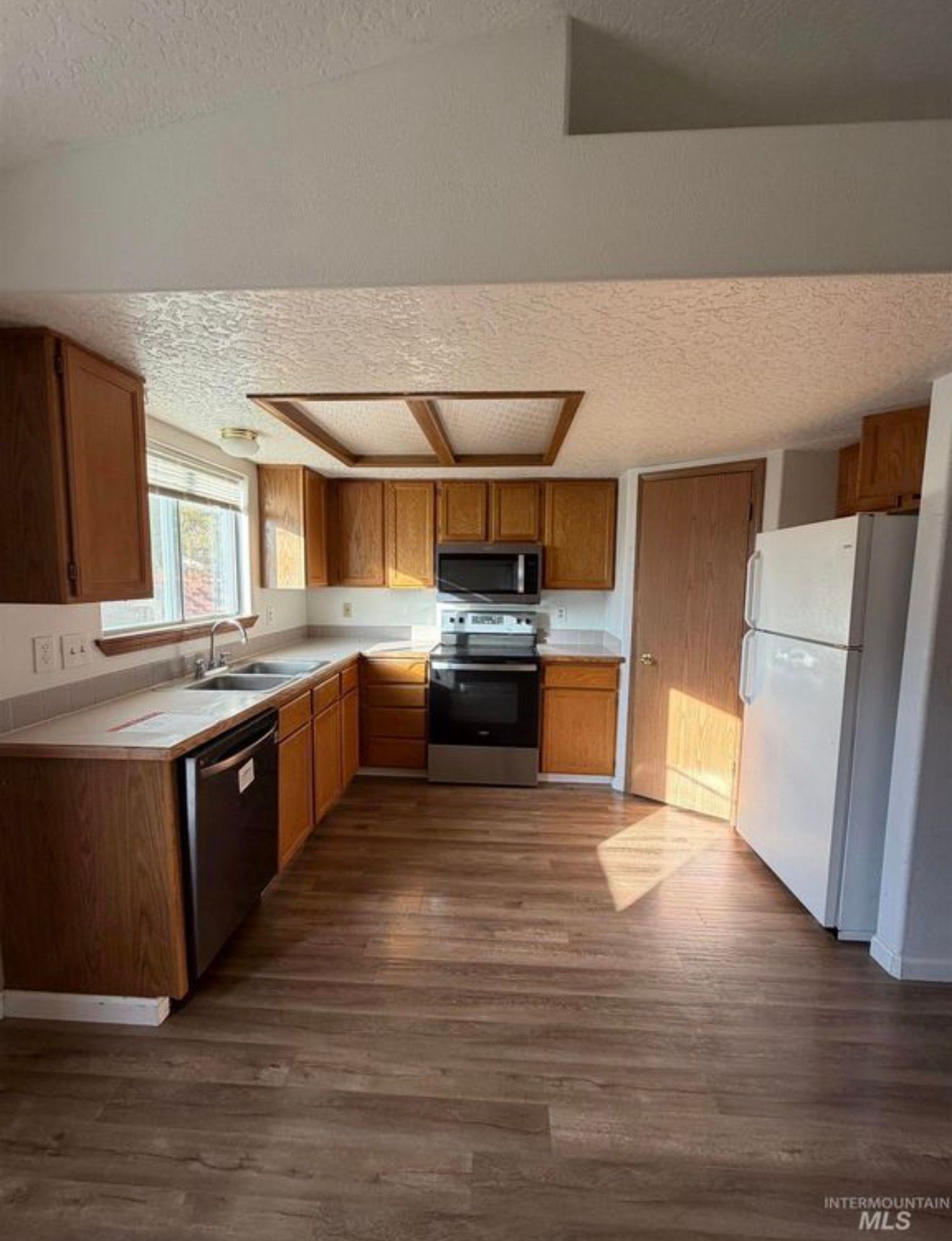 Empty kitchen with wood cabinets, white appliances, and laminate flooring, lit by a window and ceiling light.