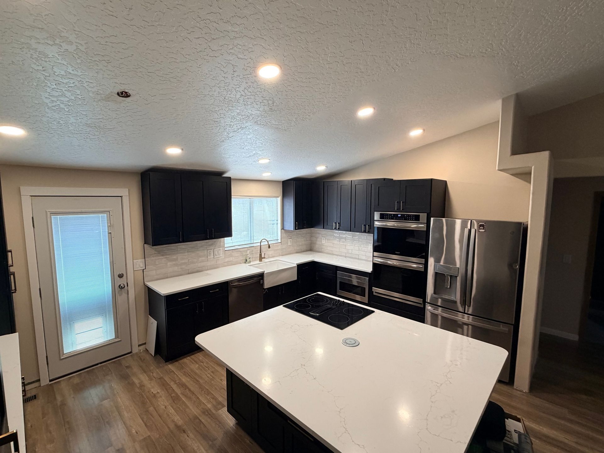 Modern kitchen with white island, dark cabinets, stainless fridge, and wood floors.