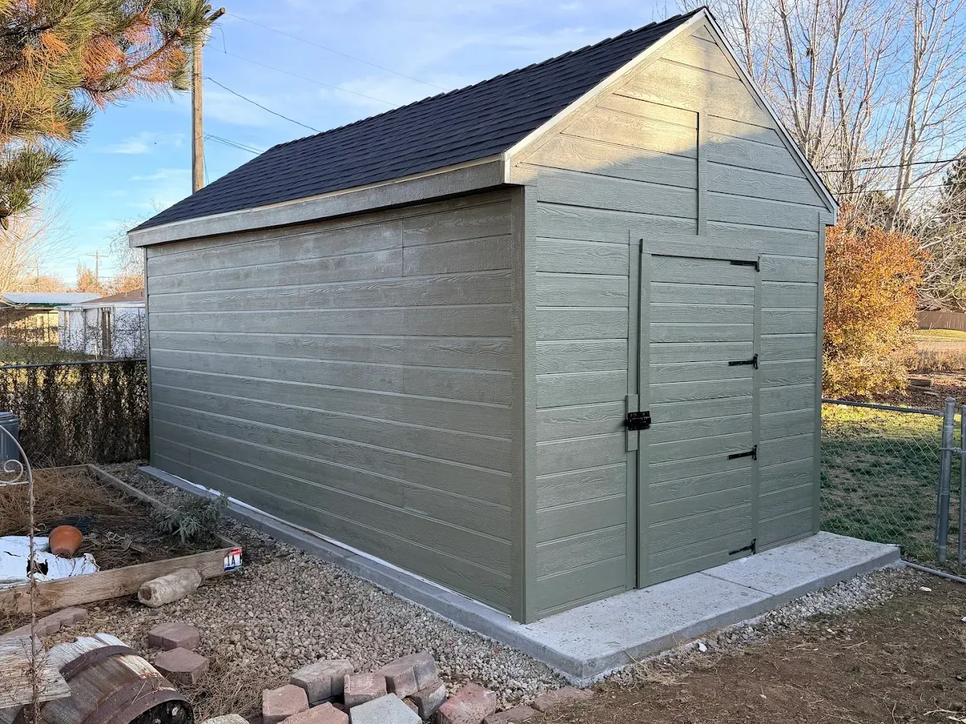 Small green backyard shed with a black roof on a concrete pad, surrounded by rocks and autumn trees