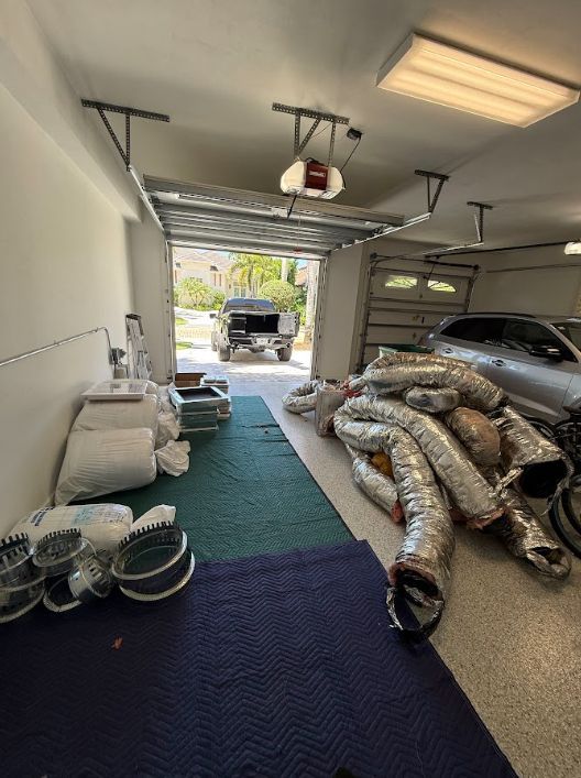 Garage filled with stacked HVAC ducts and supplies, with a car parked inside and the door open.