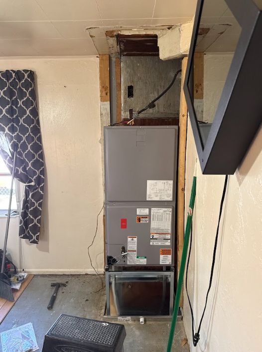 Gray furnace in a small utility room with exposed ductwork and a curtain on the left.