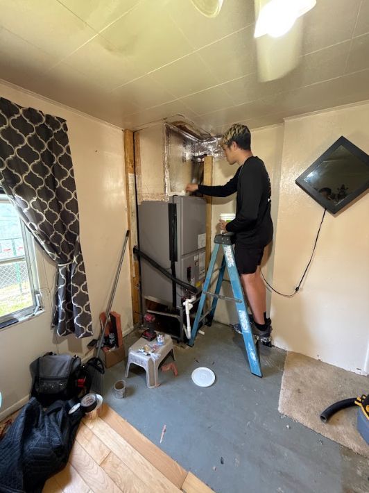 Person standing on a ladder installing ductwork in a small room with a furnace and tools on the floor