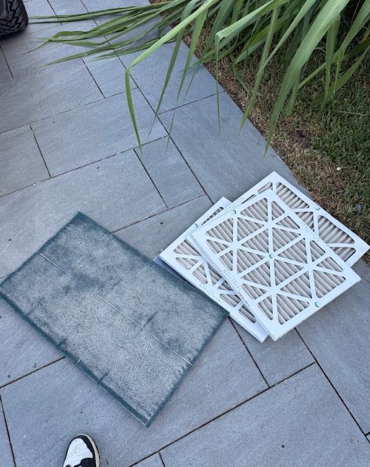 Two floor vent covers on a gray patio: one metal grate and one white plastic grate near grass.