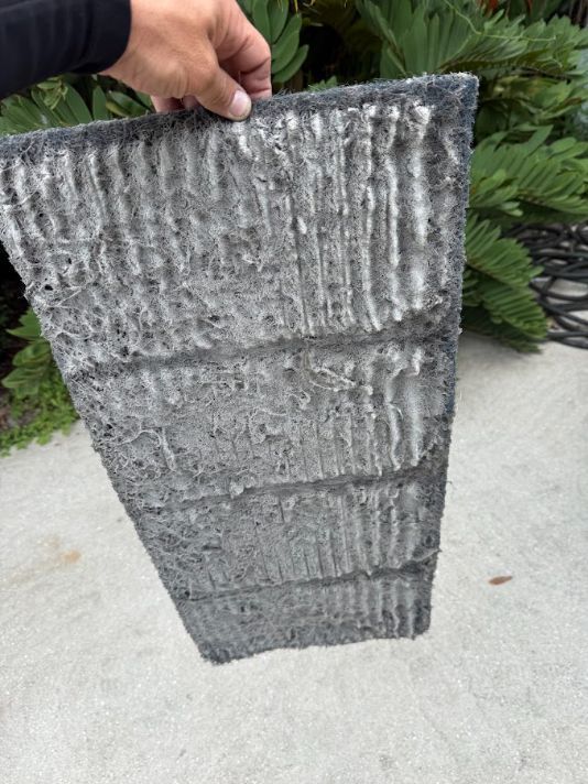 Hand holding a gray corrugated metal panel outdoors on concrete near green plants