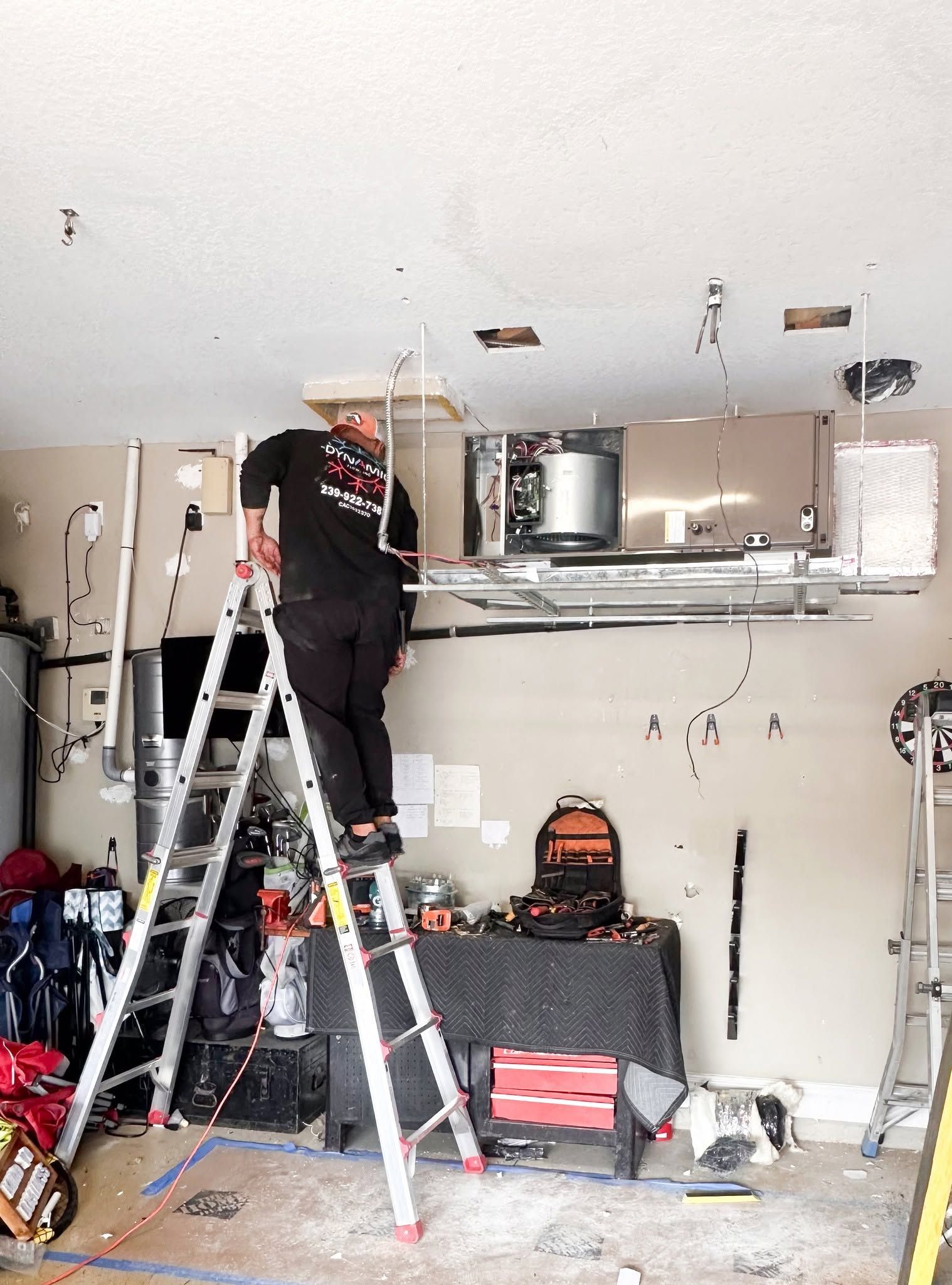 Worker on ladder installing drywall in a partially renovated room with exposed ceiling and tools below