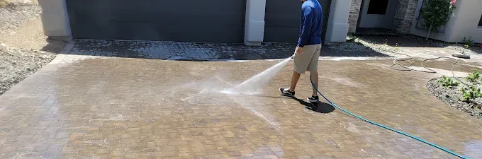 A person in a blue shirt and khaki shorts uses a pressure washer to clean a concrete driveway in front of a house.