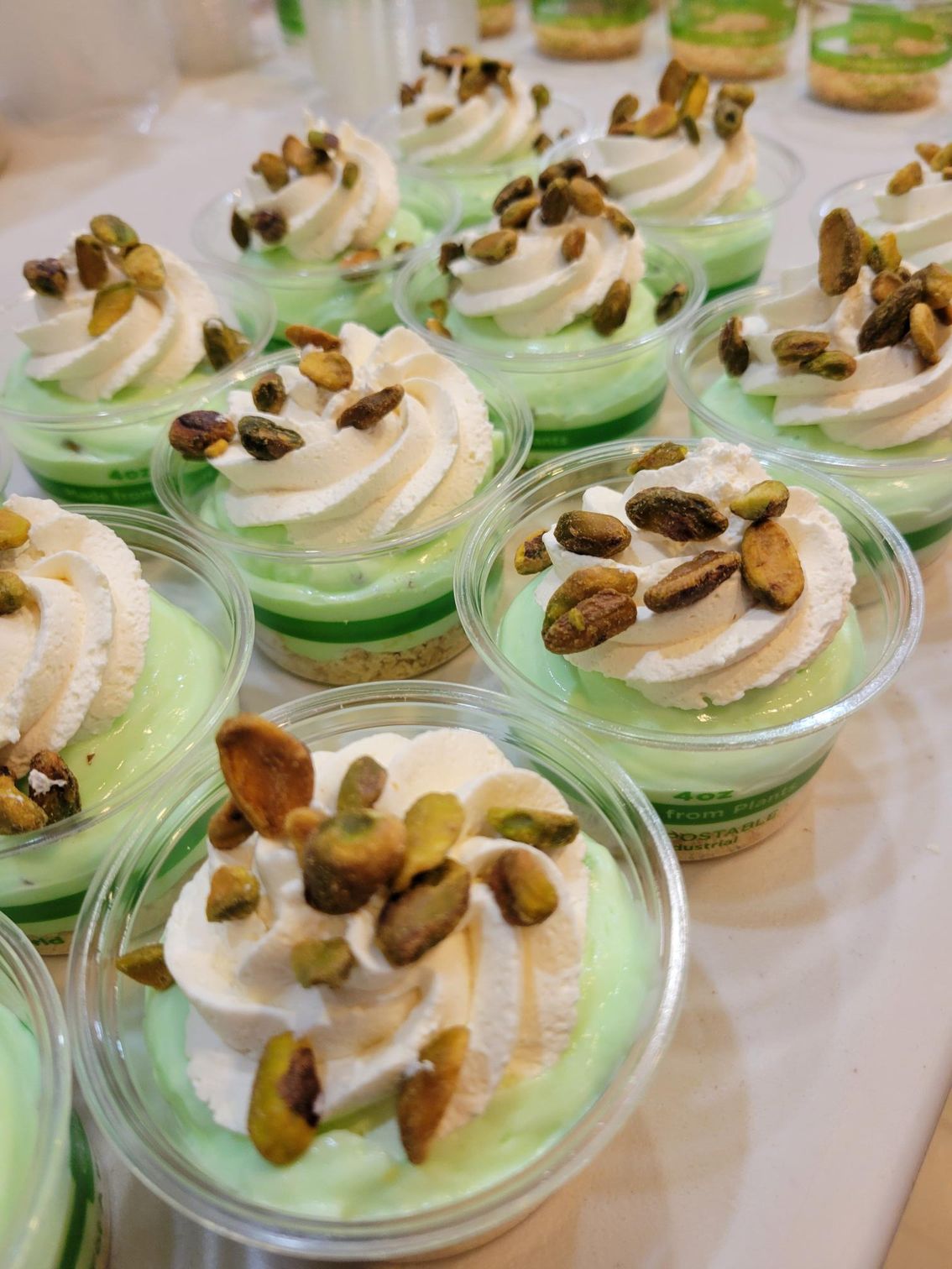 Mini dessert cups with green filling, swirled cream, and pistachio toppings arranged on a table.