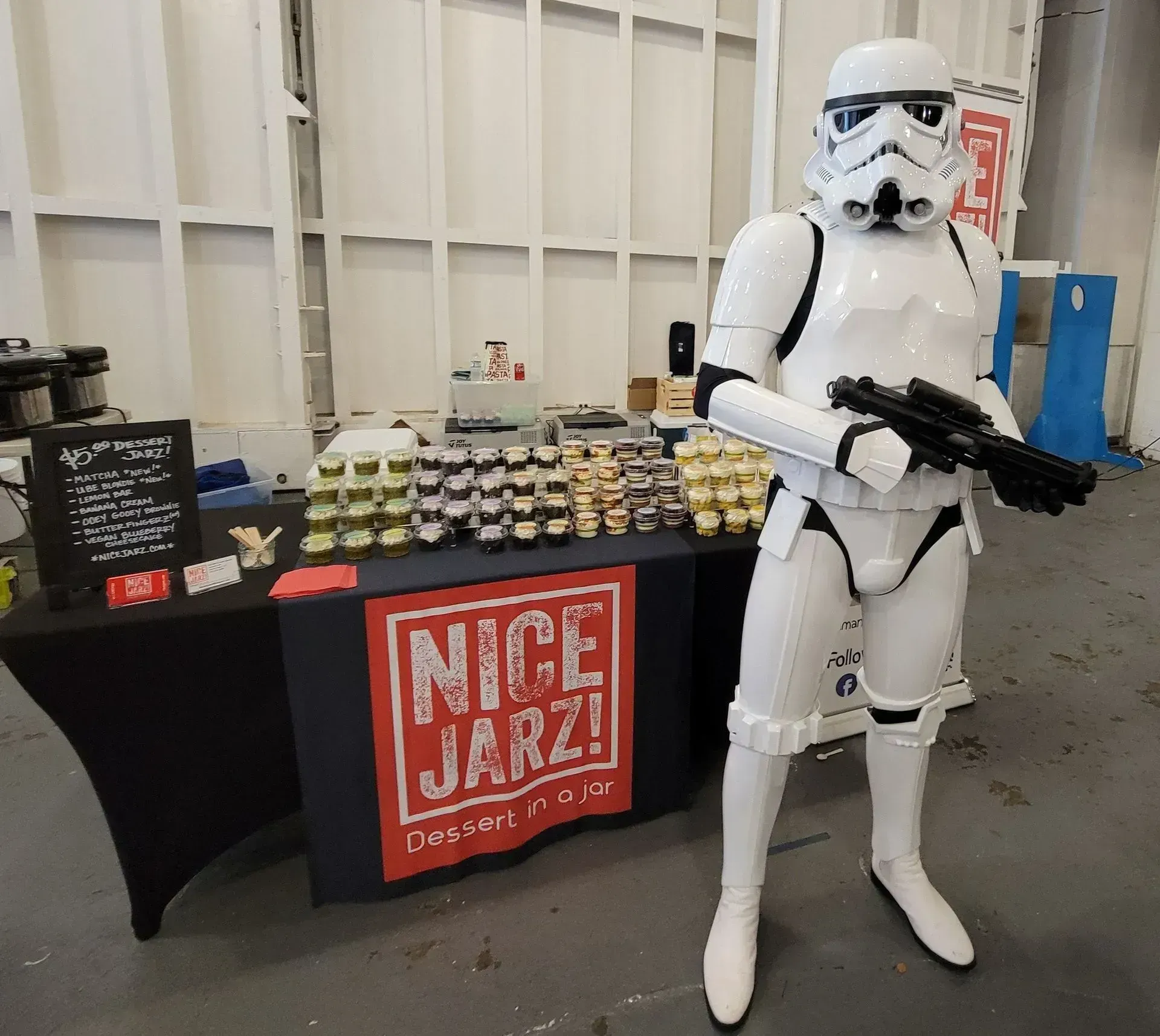 Stormtrooper costume beside a dessert table with a red “Nice Jars” sign in an indoor hall