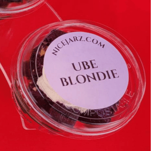 Clear plastic dessert cup labeled “UBE BLONDIE” on a bright red surface.