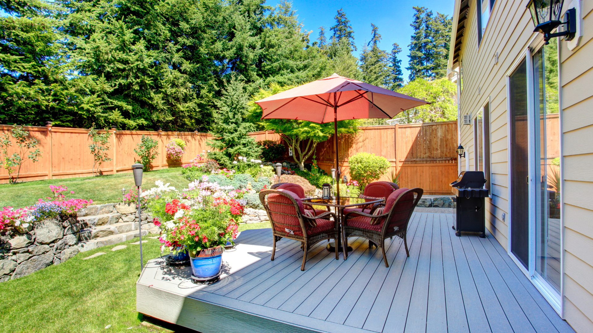 Patio with table, red umbrella, potted flowers, and a deck beside a house on a sunny day