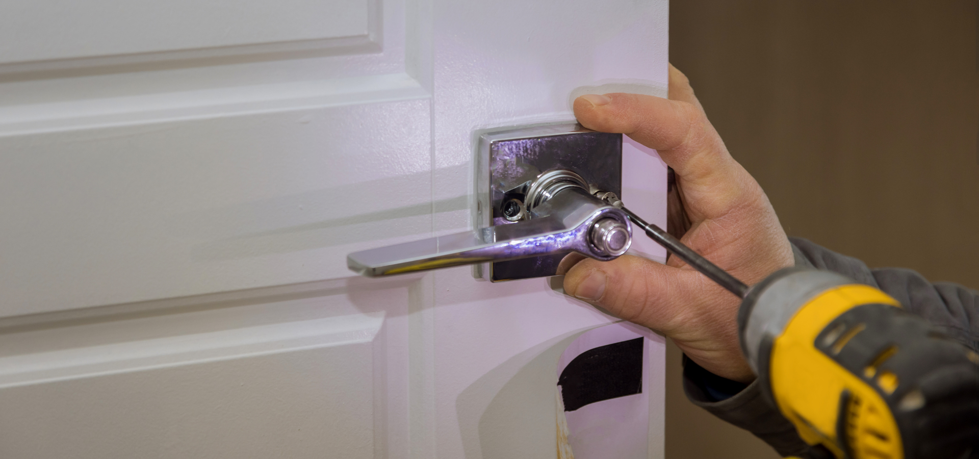 A hand using a power drill to install or secure a square, chrome-finished door handle onto a white door.