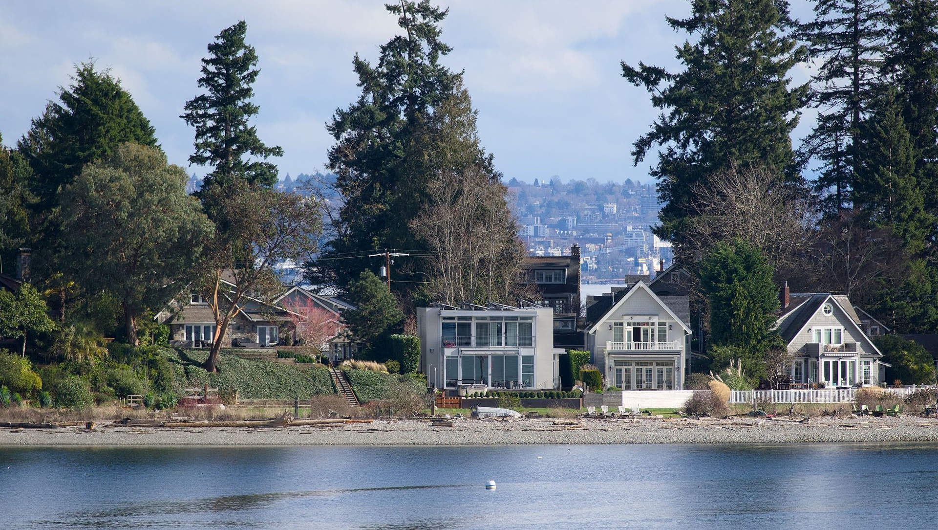 Lakeside homes with tall evergreen trees reflected in calm water under a clear sky.