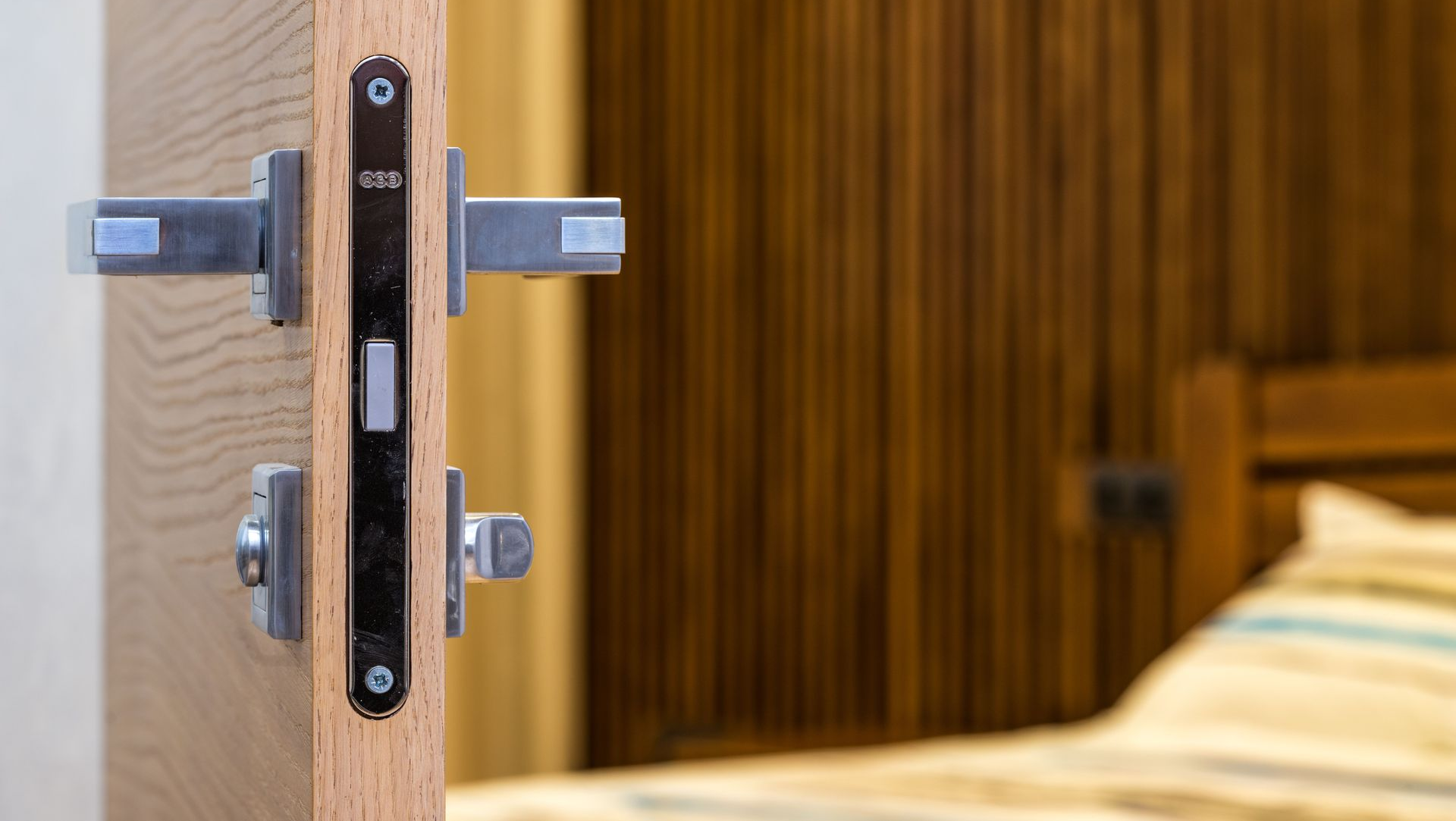Close-up of a door latch and strike plate on a wooden door, with a bedroom visible in the background
