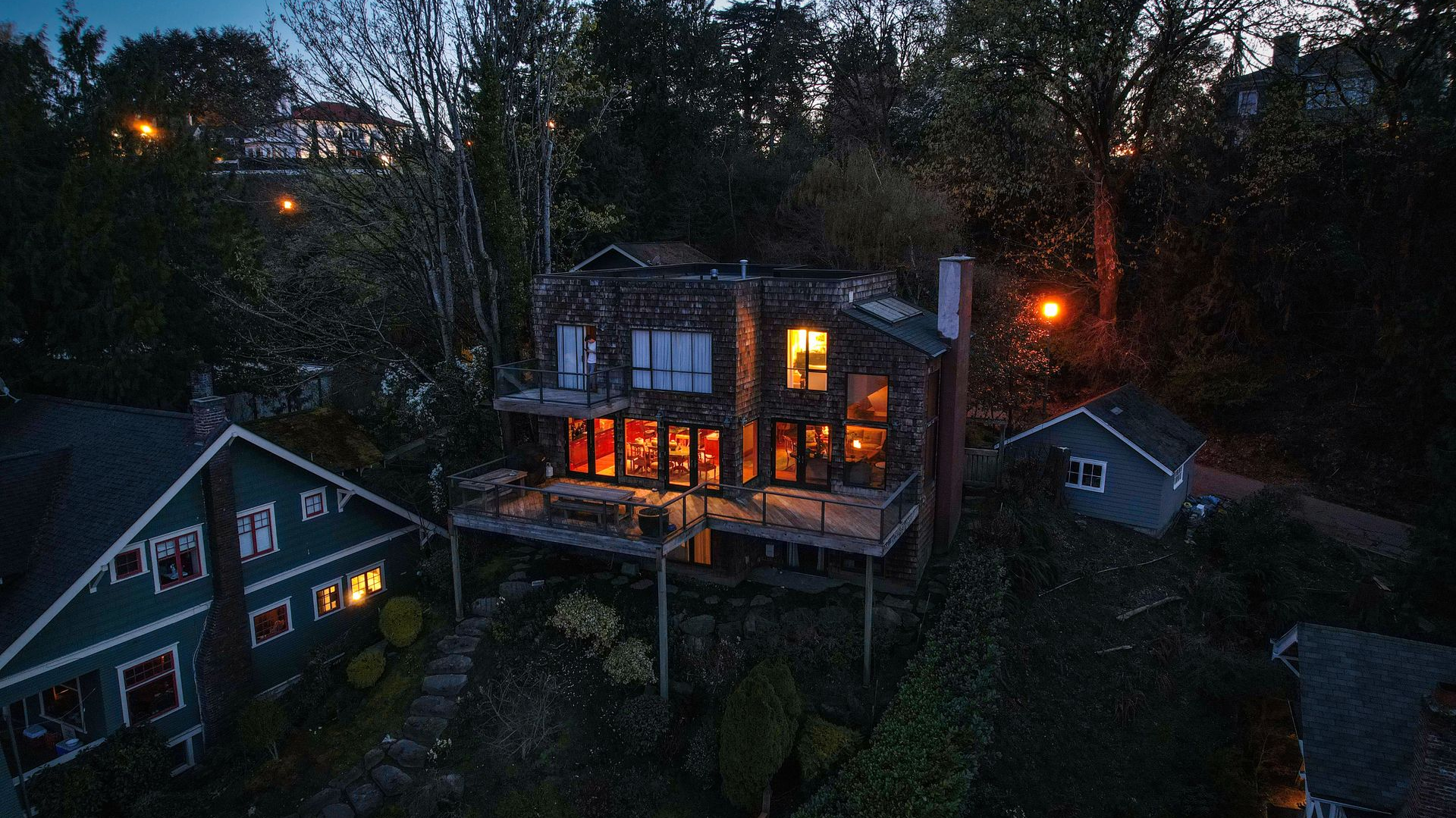 Dusk view of a lit house among trees and neighboring homes, with warm windows glowing in the dark.