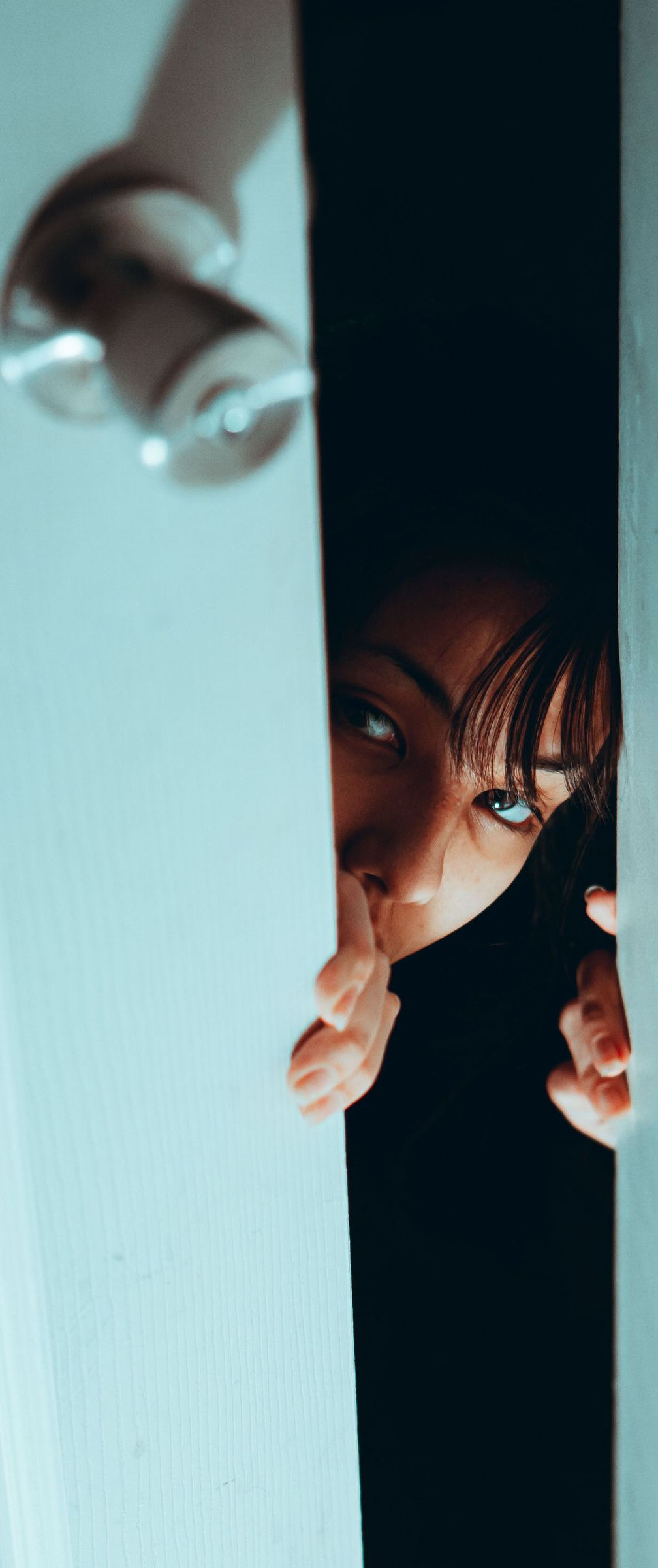 Person peeks through a narrow door gap, holding the door frame with a wary look.