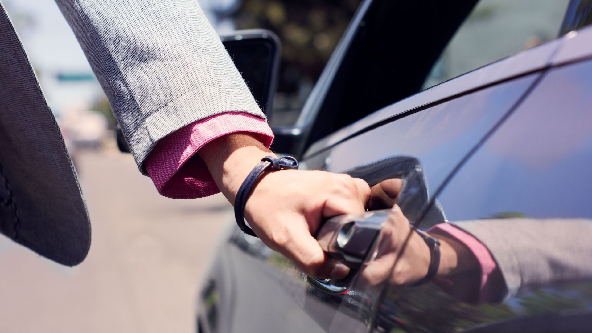 Hand reaching to open a car door, with a gray sleeve and pink cuff reflected in the window.