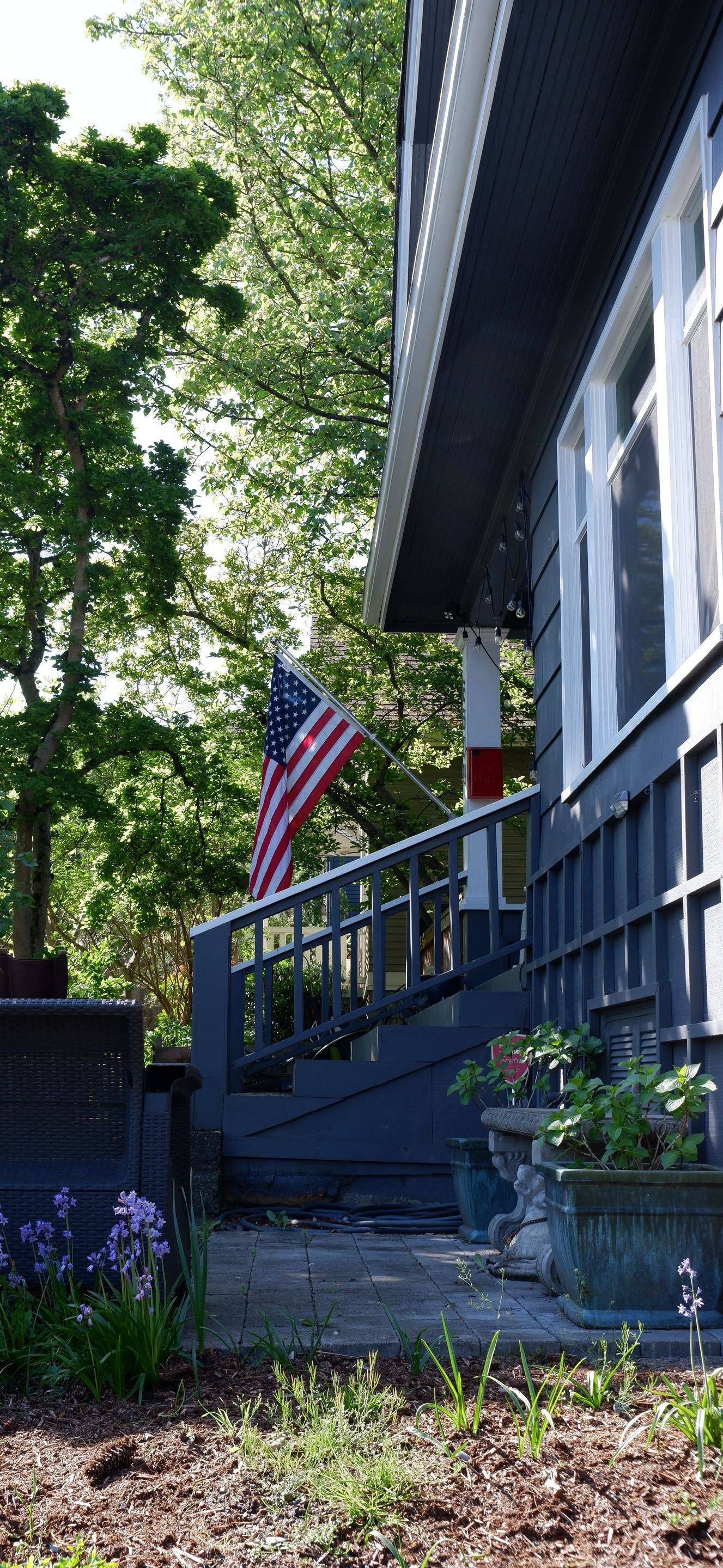 House front porch with blue railing, American flag, and flowers in a leafy yard