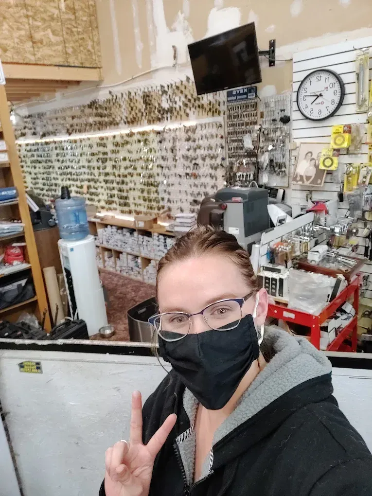 Woman wearing a mask gives a peace sign in a key shop. Keys and tools are visible in the background.