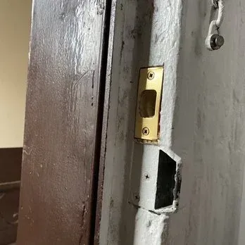 Brown door frame with a brass strike plate on a white wall. The wall has a large hole below the strike plate.