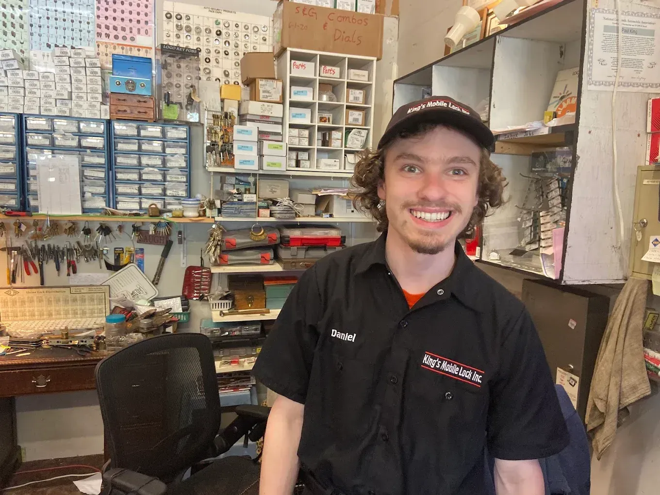 Man smiling, wearing a black work shirt, inside a hardware store. Shelves of tools and parts in the background.