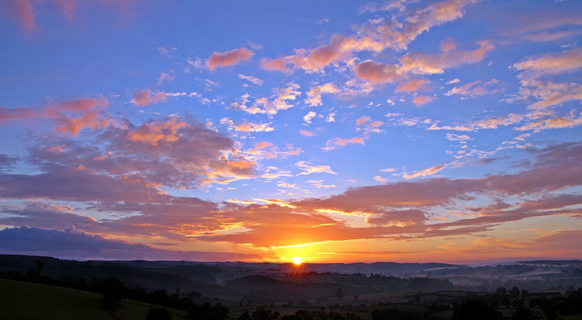 Sunrise. A new dawn breaking over the green trees and fields of the countryside. Some clouds are in the sky which is coloured with oranges, pinks, and blues.
