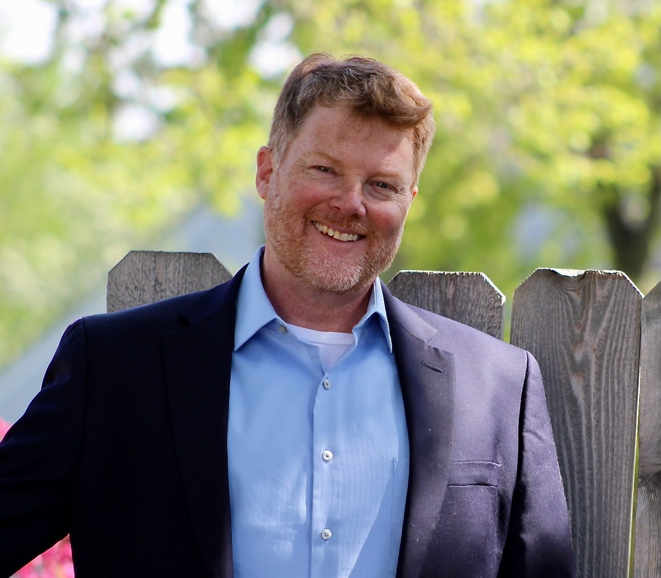 Man in blue shirt and blazer smiles by a wooden fence.