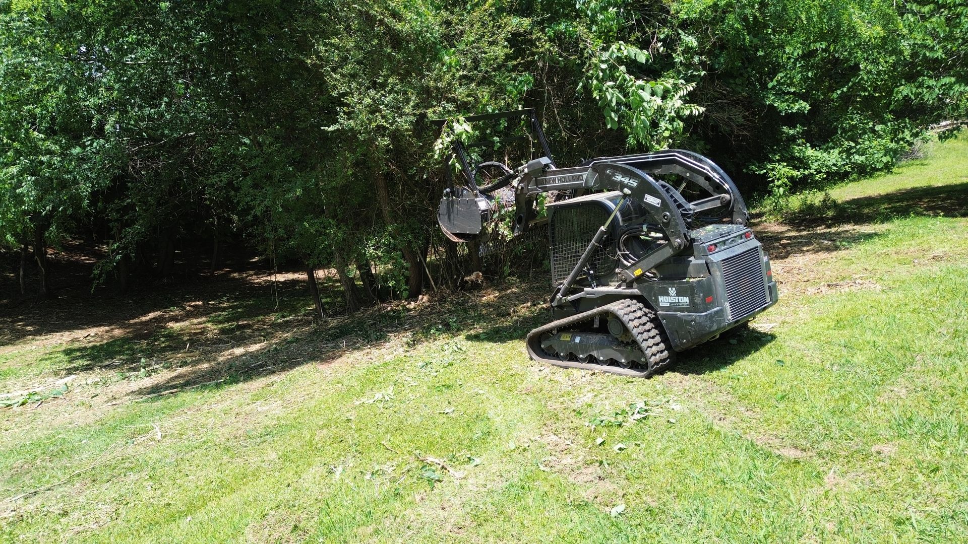 Compact tracked loader mowing grass on a sunny lawn near trees