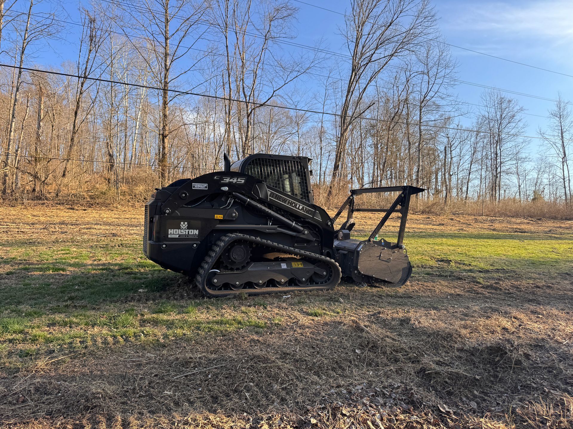 Black tracked skid-steer loader with bucket on a dirt field at the edge of a wooded area.